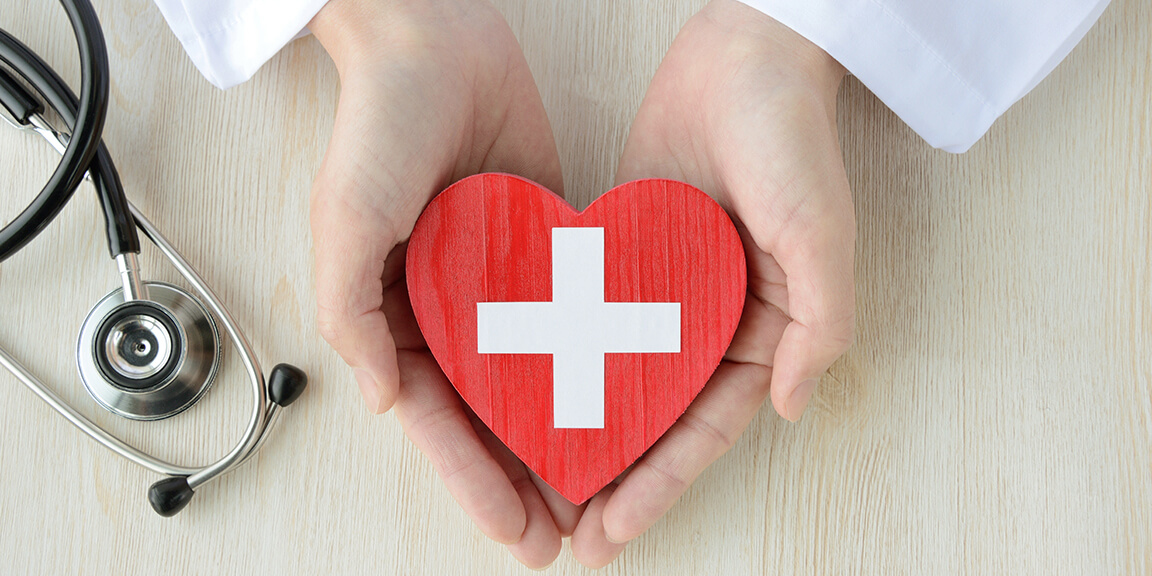 白十字が書かれたハートの模型を両手で持つ人と聴診器のイメージ